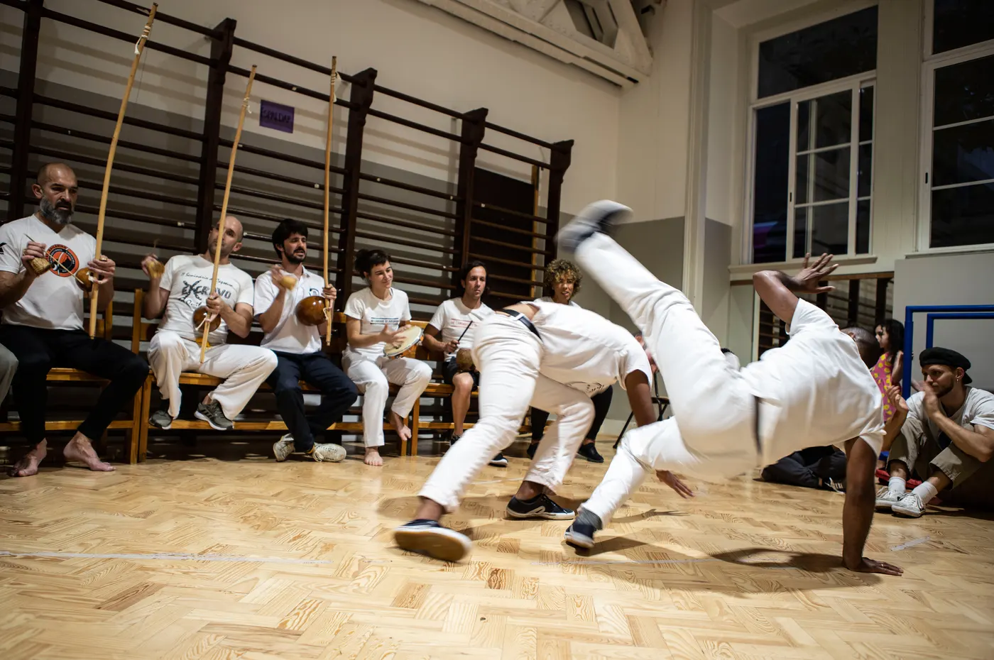 Roda de capoeira no ginásio da Academia de Capoeira de Lisboa, com dois capoeiristas a jogar e a bateria ao fundo.