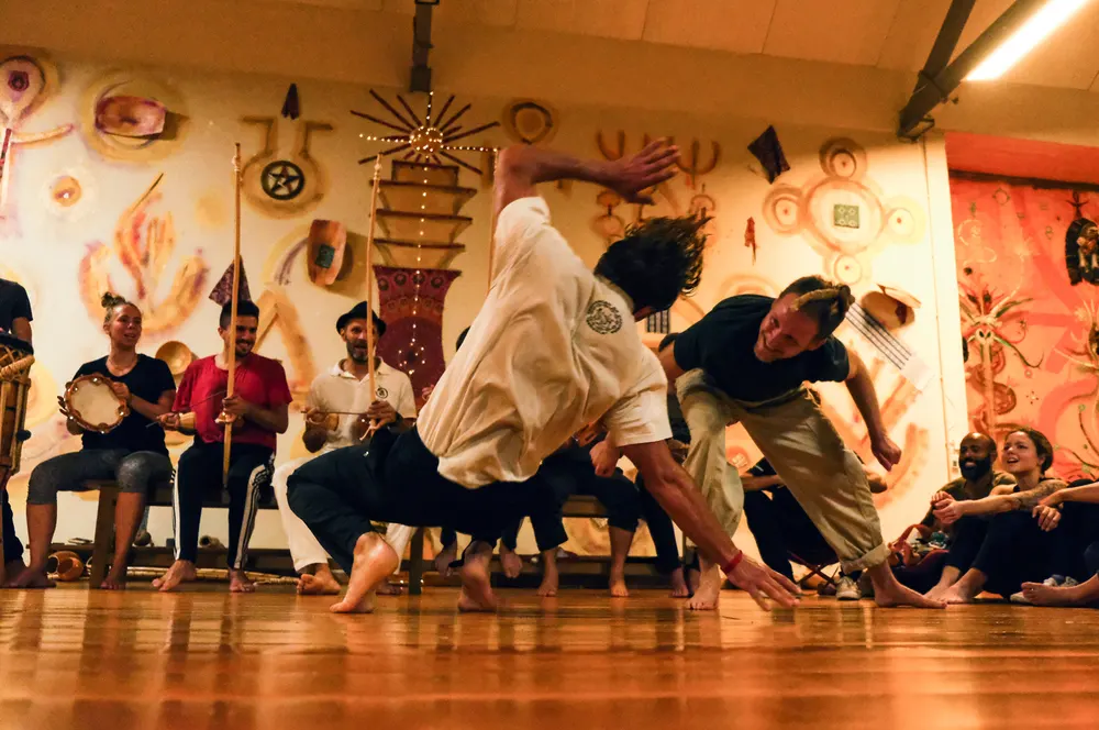 Roda de capoeira num salão pintado, dois capoeiristas a jogar junto à bateria.