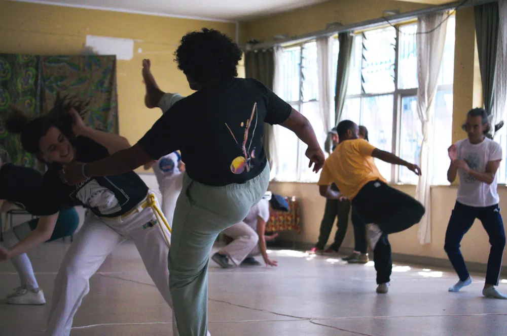 Jogo de capoeira num estúdio com janelas grandes, roda à volta.
