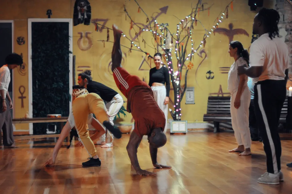 Capoeirista em bananeira numa sala com murais coloridos.